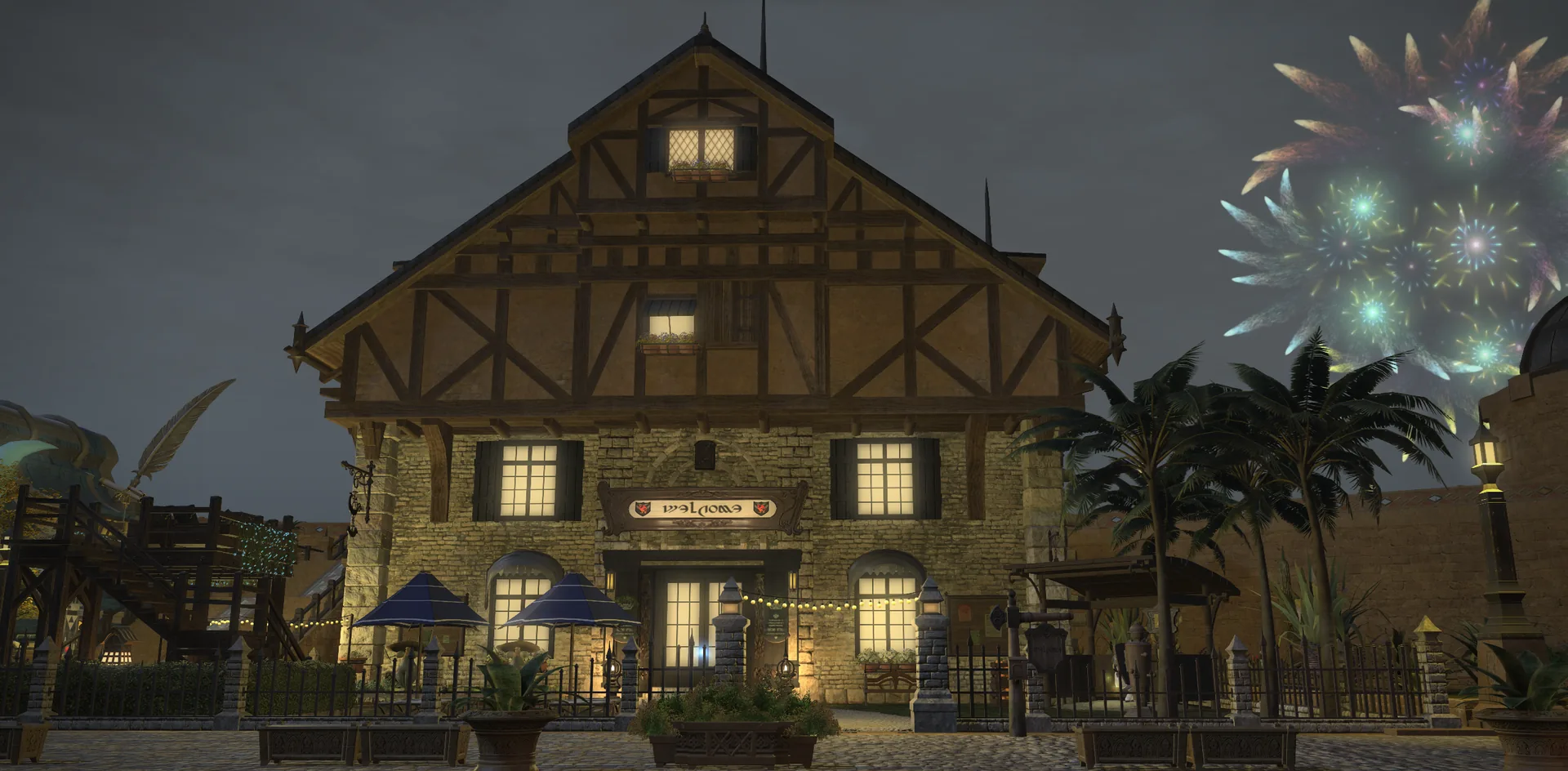 The house seen head-on at night from the cobbled street: a stone ground floor under a tall half-timbered gable, warm light in every window, a Welcome sign hung above the front doors, blue parasols and string lights over the front terrace, palm trees to either side, and fireworks bursting green and gold in the sky above the rooftops.