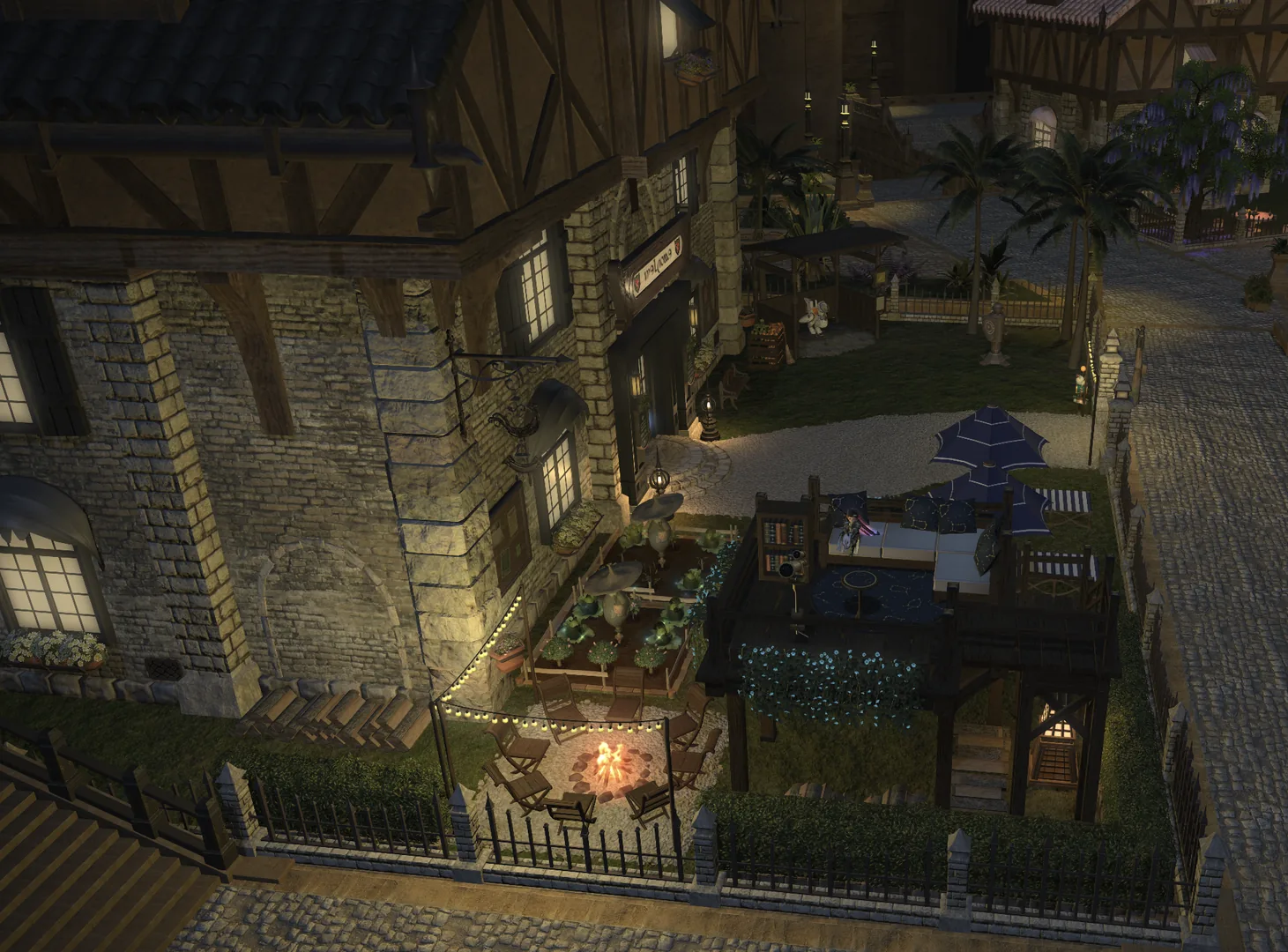 The side of the plot looked down on from above: lit windows set into the stone-and-timber wall, a raised wooden deck with a blue parasol and white seating, a firepit ringed by folding chairs on the gravel below, string lights strung between the fence posts, a garden bed of leafy greens, and hedges and palms running down to the cobbled street.